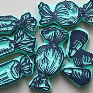 Puede incluir: Un conjunto de siete sellos de goma con forma de caramelos. Los sellos son de color turquesa con diseños en tinta negra. Los diseños presentan ilustraciones detalladas de caramelos envueltos con lazos. Ideales para manualidades.