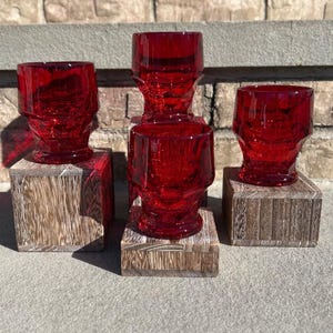 May include: Four vintage red glass goblets with a crackle effect, displayed on small wooden blocks. The goblets have a unique, geometric design and are arranged on a stone surface against a brick background. The red color is vibrant and eye-catching.