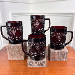 May include: A set of four deep red glass mugs featuring a textured diamond pattern on the upper section and a leaf-like motif on the lower portion. Each mug has a sturdy handle and a smooth rim, showcasing a consistent dark ruby color throughout.