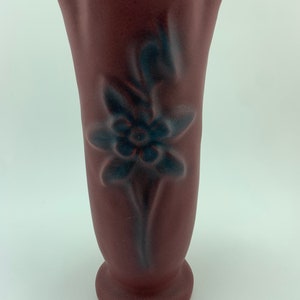 May include: A burgundy ceramic vase with a raised floral design in a darker shade of blue. The vase has a flared top and a rounded base.