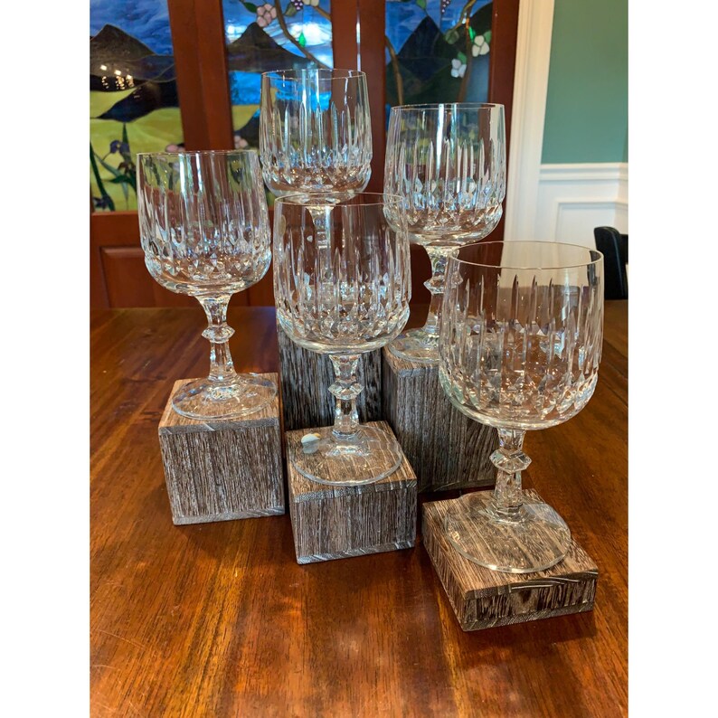 May include: Five clear crystal wine glasses with a cut glass design are displayed on individual wooden bases. The glasses are arranged in a semi-circle on a wooden table.