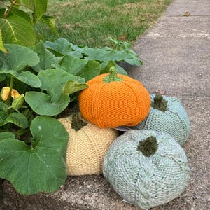 May include: Four knitted pumpkins in shades of orange, yellow, and green. The pumpkins have green stems and are arranged on a stone surface. The pumpkins are a fall-themed decorative item.