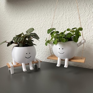 Könnte beinhalten: Zwei weiße Keramik-Pflanztöpfe in Form von lächelnden Gesichtern, einer mit einer Pflanze auf einer Holzschaukel und der andere auf einer kleinen Holzbank.