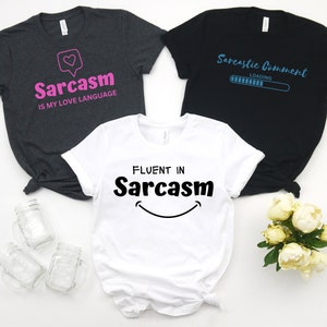 Könnte beinhalten: Drei T-Shirts mit verschiedenen sarkastischen Sprüchen. Das erste Shirt ist dunkelgrau mit pinkfarbenem Text, der "Sarkasmus ist meine Liebessprache" lautet. Das zweite Shirt ist schwarz mit blauem Text, der "Sarkastischer Kommentar wird geladen" lautet. Das dritte Shirt ist weiß mit schwarzem Text, der "Fließend in Sarkasmus" lautet, mit einem Smiley.