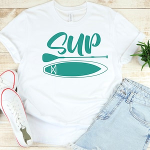 May include: White t-shirt with the word "SUP" in teal green lettering and a teal green paddleboard and paddle graphic.