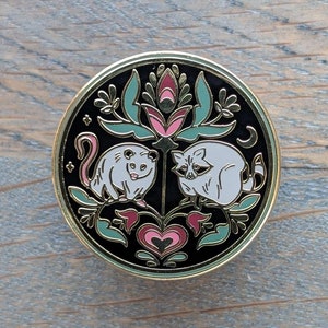 May include: A black enamel pin with a gold outline featuring a white opossum and raccoon surrounded by a floral design with pink, green, and gold accents.