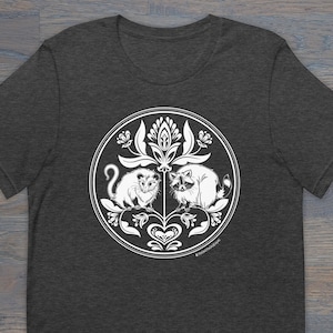 May include: A dark heather grey t-shirt with a white graphic design of a raccoon and opossum in a circle with floral designs. The text "@THISTLEFINCHDESIGNS" is at the bottom of the design.