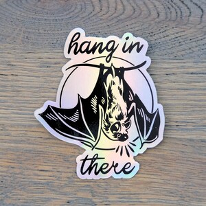 Op de afbeelding: Een holografische sticker met een zwarte vleermuis die ondersteboven hangt en de tekst "hang in there".