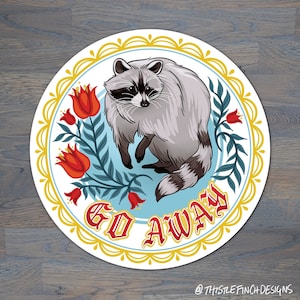 May include: A round sticker with a blue background and a white border. The sticker features a cartoon raccoon with a red, white, and blue floral design. The text "GO AWAY" is written in red letters.