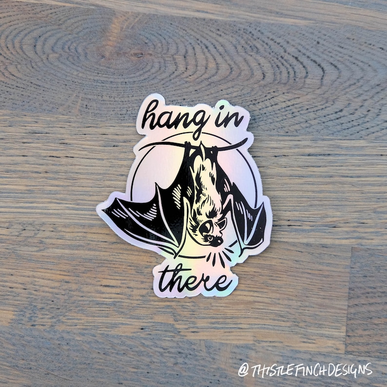 Op de afbeelding: Een holografische sticker met een zwarte vleermuis die ondersteboven hangt en de tekst "hang in there".