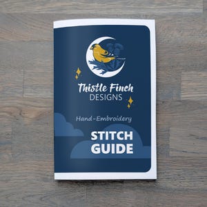 Puede incluir: Un folleto de guía de puntadas con una cubierta azul marino que presenta un diseño de pájaro amarillo y cardo dentro de una luna creciente. El texto dice "Thistle Finch Designs" y "Stitch Guide".