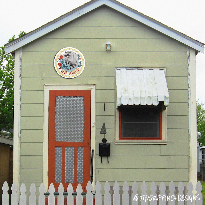 May include: A small, light green house with a white picket fence. The house has a red door with a screen, a white awning over a window, and a round sign with a pig and the words "Go Away" on it.