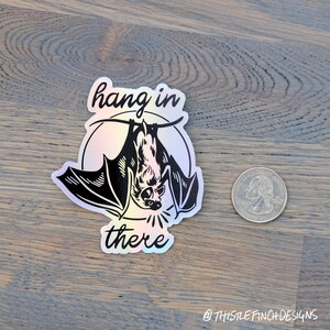 Op de afbeelding: Een holografische sticker met een zwart-witte illustratie van een vleermuis die ondersteboven hangt met de tekst "hang in there".