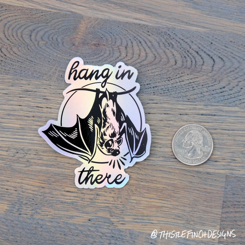 Op de afbeelding: Een holografische sticker met een zwart-witte illustratie van een vleermuis die ondersteboven hangt met de tekst "hang in there".