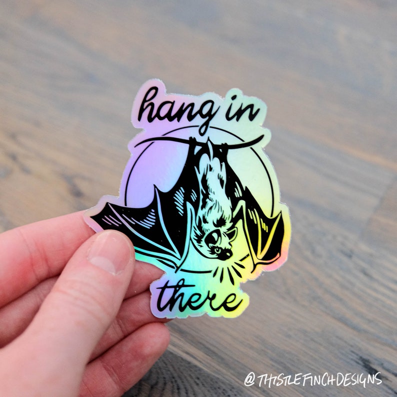 Op de afbeelding: Een holografische sticker met een zwarte vleermuis die ondersteboven hangt en de tekst "hang in there".