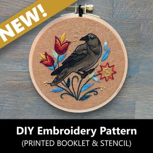 May include: Round embroidery hoop featuring a detailed embroidered design of a crow perched on a branch with flowers. The design includes red, yellow, and blue thread on a tan fabric. The text "NEW!" is in a gold banner. The text "DIY Embroidery Pattern" is below.