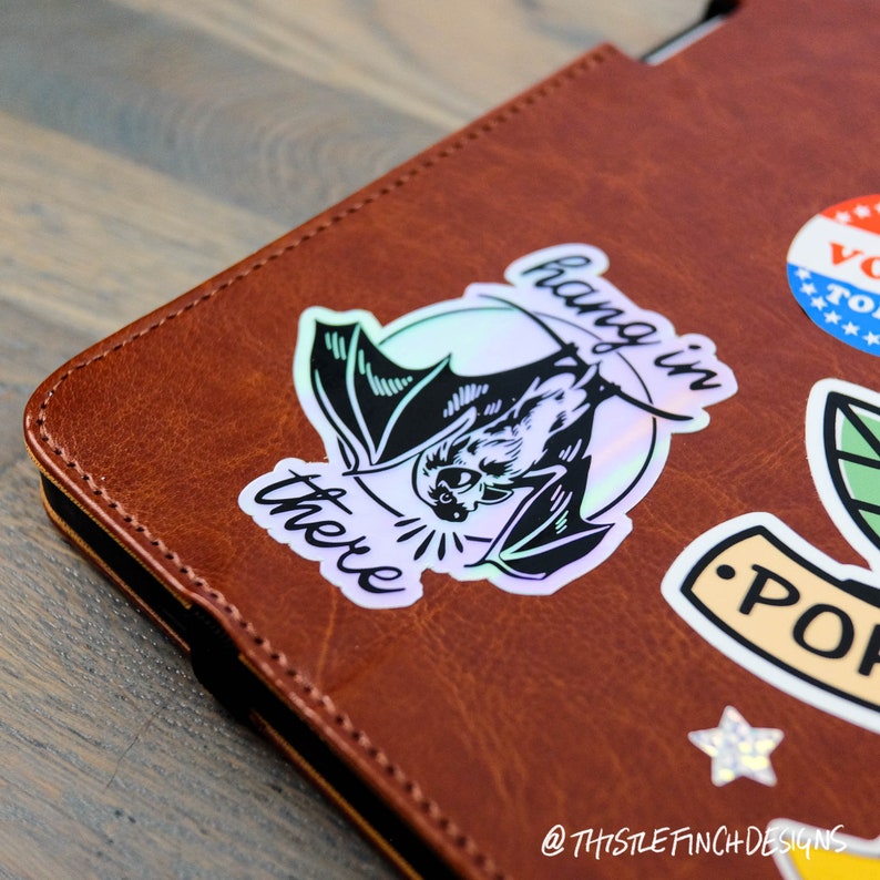 Op de afbeelding: Een holografische sticker met een zwarte vleermuis en de tekst "hang in there" op een bruin lederen oppervlak.