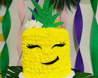 May include: A yellow pineapple-shaped cake with a green glitter pineapple topper and white flowers. The cake has a smiling face with black eyelashes and a black smile.