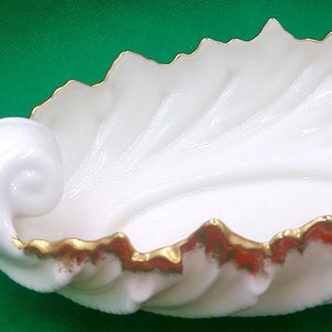 May include: White porcelain serving dish with a gold rim and a leaf-like design. The dish has a curled handle.
