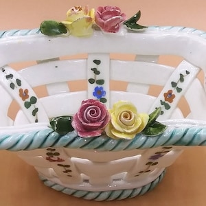 Puede incluir: Cesta de cerámica blanca con diseño tejido, adornada con detalles florales coloridos. El borde está adornado con un diseño en forma de cuerda azul claro. Presenta rosas rosas y amarillas y hojas verdes. Una pieza decorativa.