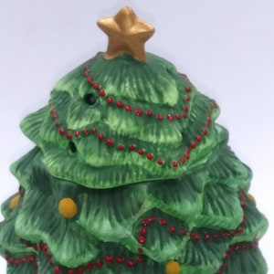 May include: A ceramic Christmas tree-shaped candle holder. The tree is green with red bead garland and gold star topper. The base is off-white with green holly leaf accents and circular cutouts.