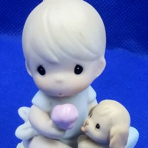 May include: A porcelain figurine of a boy holding a pink flower and a puppy sitting next to him. The boy is wearing a blue shirt and blue pants. The puppy is brown and white. The figurine is sitting on a green base with yellow flowers.