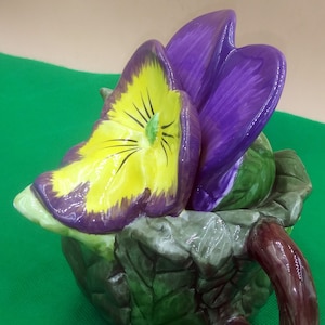 May include: A ceramic teapot shaped like a green leaf with a purple and yellow flower on top. The teapot has a brown handle.