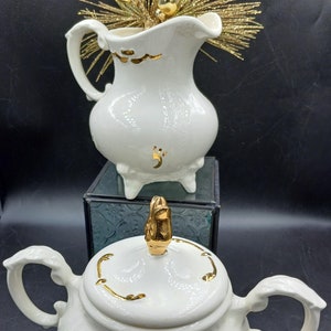 May include: White ceramic creamer and sugar set with gold accents. The creamer has a footed base and a curved spout. The sugar bowl has a lid with a gold finial and a footed base.