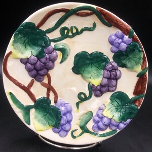 Może przedstawiać: Okrągły, kremowy ceramiczny talerz z wypukłym wzorem winogron. Winogrona są fioletowe, z zielonymi liśćmi i brązowymi pędami. Talerz jest podparty srebrnym metalowym stojakiem.
