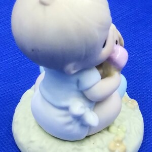 以下が含まれることがあります： ぬいぐるみを持った赤ちゃんの磁器製フィギュア。赤ちゃんは薄い青色の服を着て、花が咲いた緑色の台座に座っています。台座には「Precious Moments」という文字が刻まれています。