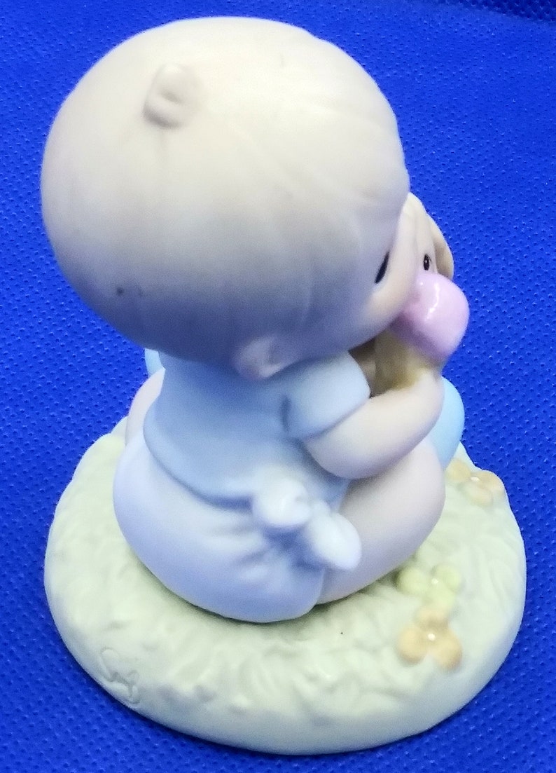 以下が含まれることがあります： ぬいぐるみを持った赤ちゃんの磁器製フィギュア。赤ちゃんは薄い青色の服を着て、花が咲いた緑色の台座に座っています。台座には「Precious Moments」という文字が刻まれています。