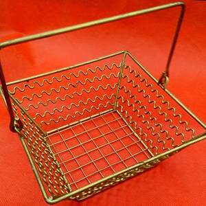 Könnte beinhalten: Ein quadratischer Metallkorb im Vintage-Stil mit Griff. Der Korb besteht aus goldfarbenem Draht mit einem wellenförmigen Muster an den Seiten und einem Gittermuster am Boden. Der Griff ist mit Scharnieren an den Seiten befestigt.