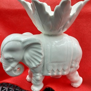 以下が含まれることがあります： 白いセラミック製の象の置物で、装飾的な花の形をした上部が付いています。象には、鼻、耳、鞍のようなデザインなど、詳細な特徴があります。置物は赤い背景に置かれています。