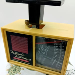 May include: A yellow plastic Weight Watchers Deluxe Food Scale with a black base and a black scale with markings in ounces and grams. The scale reads "Not Legal for Trade".