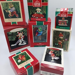 Puede incluir: Una colección de adornos Hallmark Keepsake en sus cajas originales. Los adornos presentan varios personajes, incluyendo un mapache en bicicleta, un muñeco de nieve en un coche y un castor jugando al fútbol. Las cajas son rojas con detalles verdes.