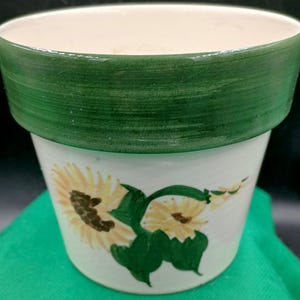 May include: A ceramic flower pot with a wide, hand-painted forest green rim and a cream-colored base. The exterior features a painted design of two yellow sunflowers with brown centers and green leaves, extending across the front of the vessel.
