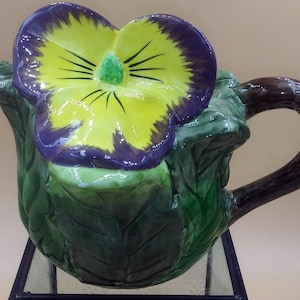 Puede incluir: Una tetera de cerámica con forma de planta verde con hojas y una flor de pensamiento púrpura y amarilla en la parte superior. La tetera tiene un asa marrón.