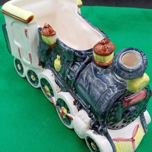 May include: A ceramic train planter in shades of blue, white, yellow, and brown. The planter is shaped like a vintage steam engine with detailed wheels and a spacious interior for plants or decorative items. The train has a classic design.