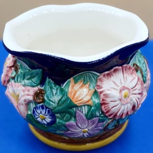 May include: A ceramic flower pot with a white interior and a dark blue rim. The exterior features a relief design of colorful flowers and green leaves. The base is yellow.