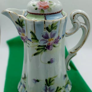 Peut inclure: Une cafetière en porcelaine blanche avec un couvercle, ornée de fleurs violettes et de feuilles vertes peintes à la main. La cafetière a une bordure et une poignée dorées, avec une rose rose sur le couvercle. Le design comprend des accents bleus et verts.