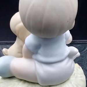 以下が含まれることがあります： 小さな犬と一緒に座っている若い男の子の磁器製フィギュア。男の子は青いシャツと白いズボンを履いています。犬は白地に茶色の斑点があります。フィギュアは「Sam」と書かれた緑色の台座の上に座っています。