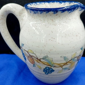 May include: Cream ceramic pitcher with a large handle and a blue rim. The pitcher is decorated with a vine design featuring blue grapes, green and yellow leaves, and brown stems. The pitcher has a speckled finish.