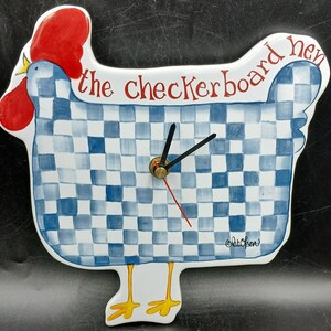 May include: A ceramic wall clock shaped like a hen with a blue and white checkerboard pattern. The hen has a bright red comb and wattle, yellow feet, and the text "the checkerboard hen" written in red. Black clock hands are centered on the body.