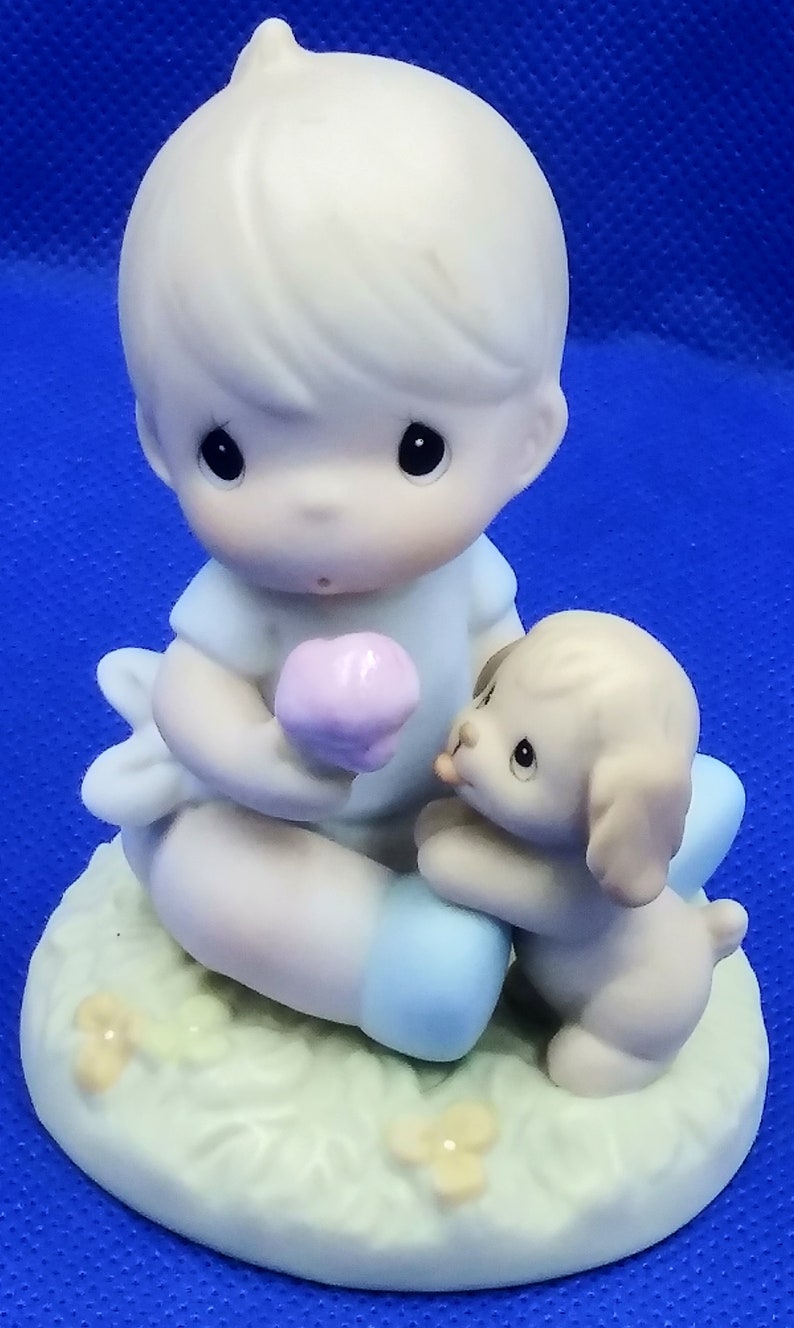 May include: A porcelain figurine of a boy holding a pink flower and a puppy sitting next to him. The boy is wearing a blue shirt and blue pants. The puppy is brown and white. The figurine is sitting on a green base with yellow flowers.