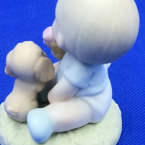 以下が含まれることがあります： 小さな子供が子犬と一緒に座っている磁器製の置物です。子供は青いシャツを着ていて、髪にピンクの花をつけています。子犬は茶色と白で、子供の隣に座っています。置物は緑色の台座の上にあります。