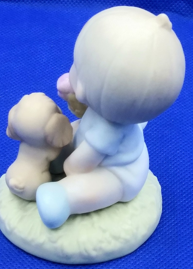 以下が含まれることがあります： 小さな子供が子犬と一緒に座っている磁器製の置物です。子供は青いシャツを着ていて、髪にピンクの花をつけています。子犬は茶色と白で、子供の隣に座っています。置物は緑色の台座の上にあります。