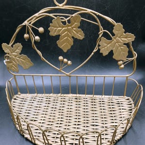 Puede incluir: Una cesta dorada para colgar en la pared con una base tejida y detalles decorativos de hojas y bayas. La cesta tiene forma curva y un marco de metal con un lazo para colgar.
