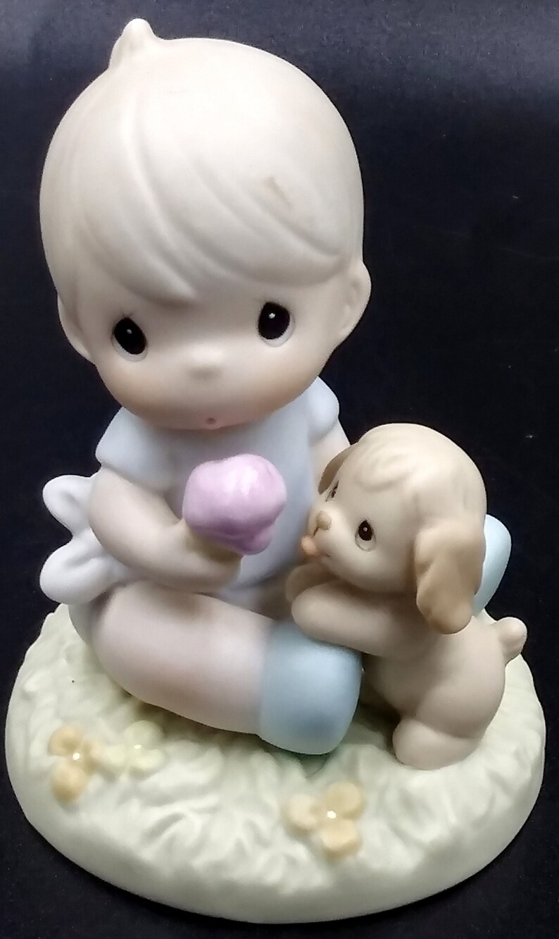 以下が含まれることがあります： 男の子がアイスクリームコーンを持って、隣に子犬が座っている、磁器製の置物です。男の子は青いシャツと白いズボンを履いています。子犬は茶色と白です。置物は緑色の土台の上に置かれ、黄色の花が飾られています。
