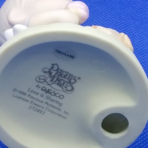 以下が含まれることがあります： ピンクの花を持ったプレシャスモーメントの天使の白い磁器の置物。置物の台座は白で、「プレシャスモーメント」と「愛は分かち合うこと」の文字が印刷されています。置物はタイ製です。