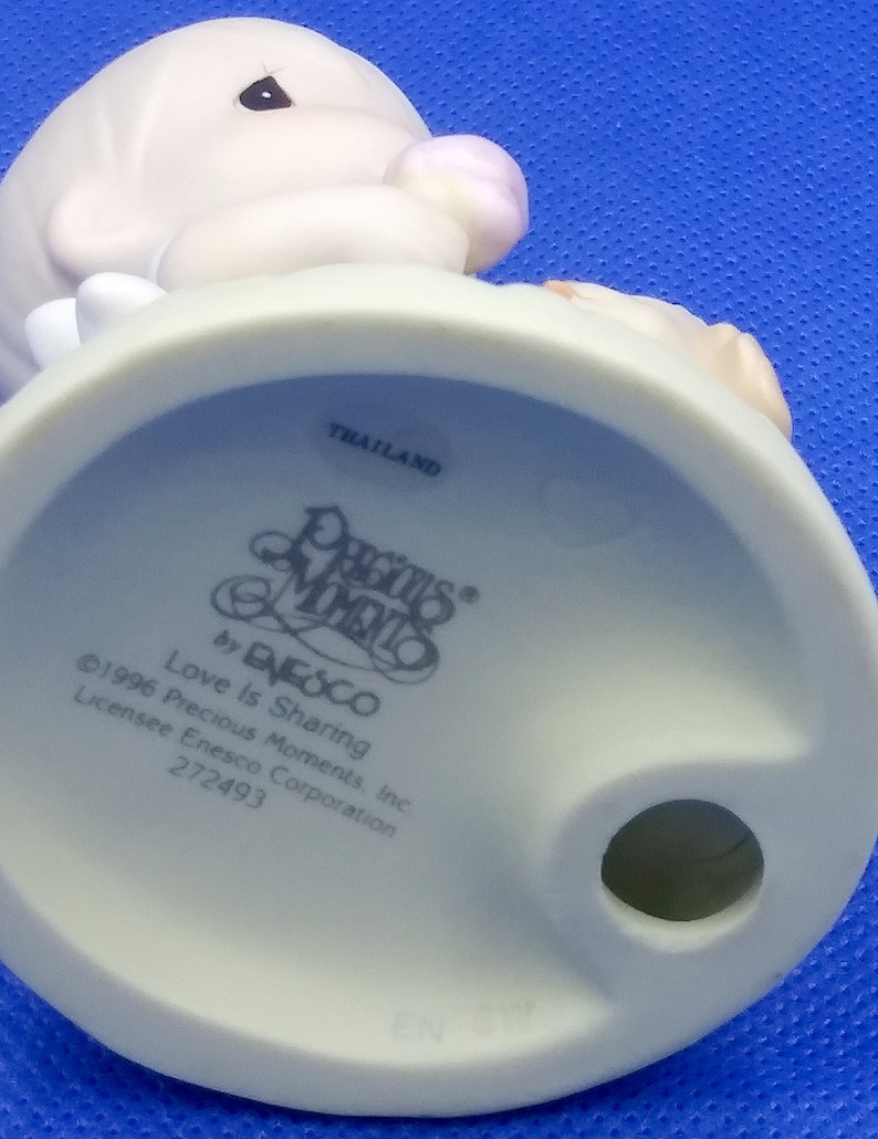 以下が含まれることがあります： ピンクの花を持ったプレシャスモーメントの天使の白い磁器の置物。置物の台座は白で、「プレシャスモーメント」と「愛は分かち合うこと」の文字が印刷されています。置物はタイ製です。